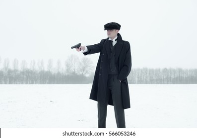 Retro 1920s english gangster with black coat and flat cap shooting with gun in winter snow landscape. Peaky blinders style.