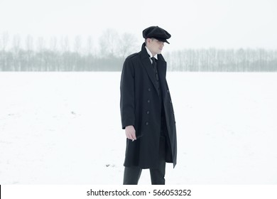 Retro 1920s english gangster with black coat and flat cap standing in winter snow landscape. Peaky blinders style.