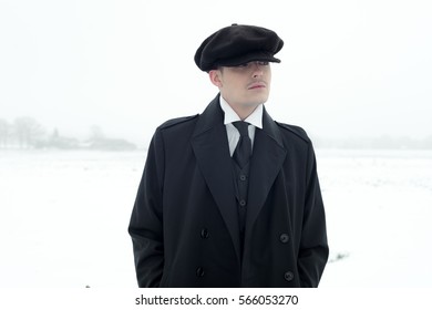 Retro 1920s english gangster with black coat and flat cap standing in winter snow landscape. Peaky blinders style.