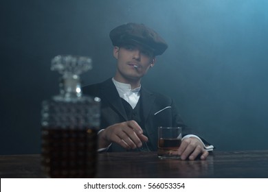 Retro 1920s english gangster sitting at table with whiskey. Peaky blinders style.