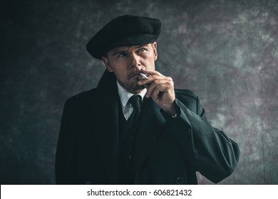 Retro 1920s english gangster smoking cigarette. Wearing black coat and flat cap.