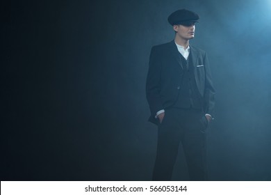 Retro 1920s english gangster wearing suit and flat cap standing in smoky room. Peaky blinders style.