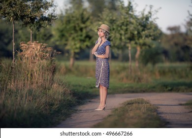Retro 1920s Summer Fashion Woman With Blue Dress And Straw Hat Standing On Rural Road.