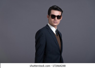 Retro 50s Business Fashion Man With Dark Grease Hair. Wearing Dark Blue Suit And Sunglasses. Studio Shot Against Grey.