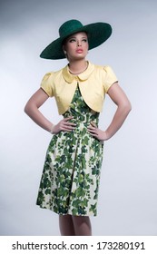 Retro 50s Fashion Brunette Girl With Hat Wearing Green Dress And Yellow Blouse. Studio Shot Against Grey.