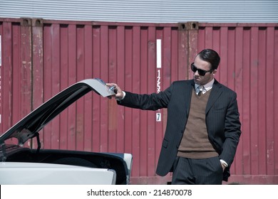 Retro 60s Mafia Business Man With Suit And Black Sunglasses Looking In Trunk Of Classic Car.