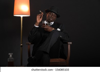 Retro african american gangster man wearing striped suit and tie and black hat. Sitting in a chair in living room. Smoking cigar. Holding glass of whisky. - Powered by Shutterstock