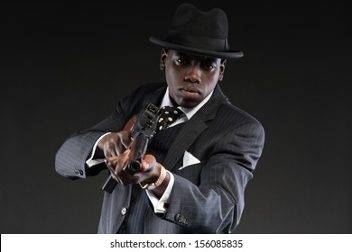 Retro African American Mafia Man Wearing Striped Suit And Tie And Black Hat. Shooting With Machine Gun. Studio Shot.