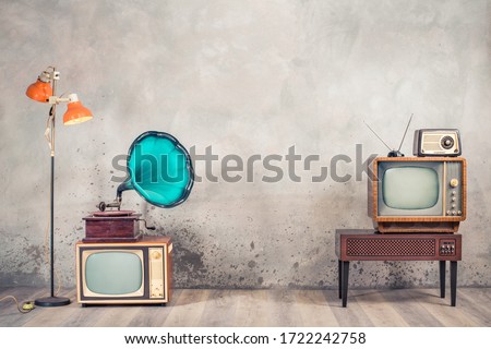 Retro analog CRT televisions, wooden TV stand with outdated amplifier, old radio receiver from 60s, classic gramophone, floor lamp front aged concrete wall background. Vintage style filtered photo