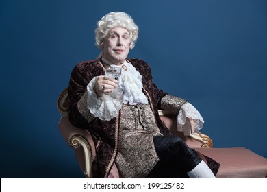 Retro Baroque Man With White Wig Holding A Wine Glass Sitting On Antique Couch. Studio Shot Against Blue.