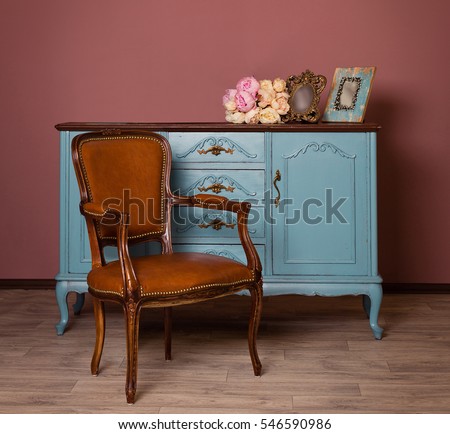 Retro brown leather armchair near blue dresser, tender bouquet and two frames. Blue and brown vintage interior. Brown room with ethnic dresser and chair. Antique cupboard. Clothes closet. Vanity Table
