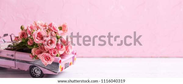Retro car toy with pink roses flowers against  pink\
textured  wall. Long bannerformat. Romantic background. Selective\
focus. Place for text.