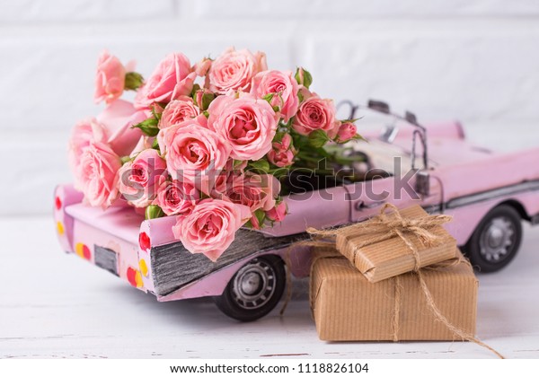 Retro car toy with pink roses and\
wrapped boxes with presents  flowers against  white textured  wall.\
Romantic background. Selective focus. Place for\
text.