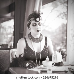 Retro Charleston Woman Sitting with in a Restaurant Holding a Cup of Tea. Black and White Portrait of The Beautiful Retro Woman Drinking Tea in Cafe in Black Lace and Accessories in Style 1920 - 1930 