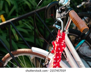Retro city bike with red closed umbrella hanging on synthetic leather handlebar grip , white stylish bicycle, green bushes on background - Shutterstock ID 2157457019