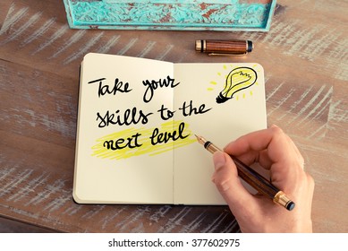 Retro Effect And Toned Image Of A Woman Hand Writing A Note With Fountain Pen On A Notebook. Handwritten Text TAKE YOUR SKILLS TO THE NEXT LEVEL Next To Yellow Lighting Bulb As Symbol For Bright Idea