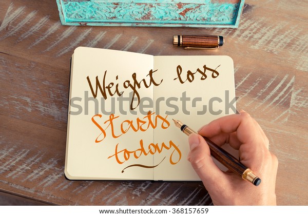 Retro effect and toned image of\
a woman hand writing a note with a fountain pen on a notebook.\
Handwritten text WEIGHT LOSS STARTS TODAY, motivation\
concept