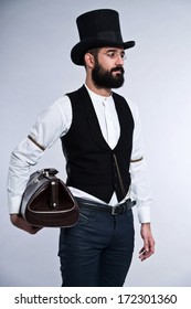 Retro hipster 1900 fashion man with black hair and beard. Wearing black hat. Holding vintage bag. Studio shot against grey.