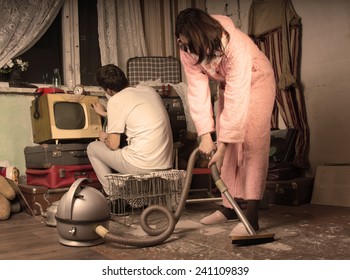 Retro Housewife In Her Dressing Gown And Slippers Cleaning A Messy Living Room With A Vintage Vacuum Cleaner While Her Husband Watches Television On An Old TV Set, Aged Style Toning