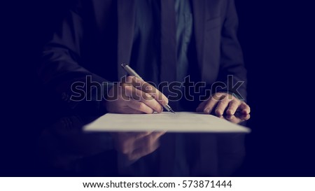 Retro image of businessman signing document or contract with silver pen on a black desk with reflection.