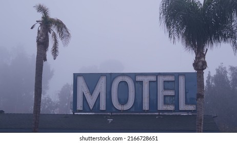 Retro neon sign of motel or hotel on road, foggy misty weather in California, USA. Palm trees in haze, gloomy moody atmosphere. Lodging on hitchhiking road trip. Tourism or traveling in America