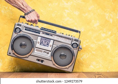 Retro outdated portable stereo boombox radio receiver with cassette recorder from circa 1980s in a strong man's hand front concrete textured yellow wall background. Vintage old style filtered photo 