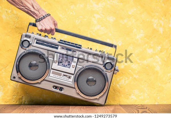 Retro outdated portable stereo boombox radio receiver\
with cassette recorder from circa 1980s in a strong man\'s hand\
front concrete textured yellow wall background. Vintage old style\
filtered photo 