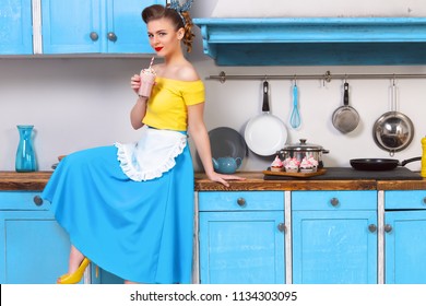 Retro pin up girl woman female housewife wearing colorful top, skirt and white apron holding cooked sweet strawberry milkshake sitting in the kitchen with utensils and tray with cupcakes.  - Powered by Shutterstock