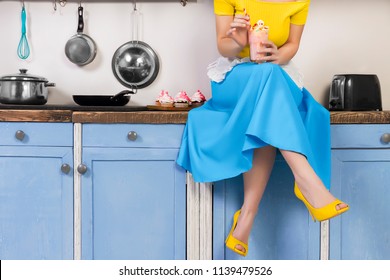 Retro pin up girl woman female housewife wearing colorful top, skirt and white apron holding cooked sweet strawberry milkshake sitting in the kitchen with utensils and tray with cupcakes. - Powered by Shutterstock