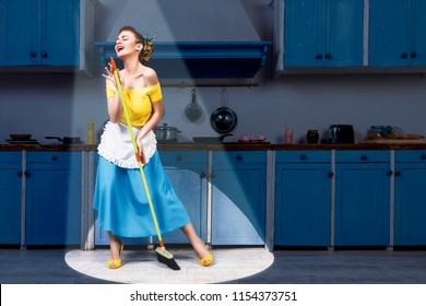 Retro pin up girl woman female housewife wearing yellow top, blue skirt and white apron holding mop singing and cleaning floor in stage light kitchen with blue cabinets and utensils. Housework concept