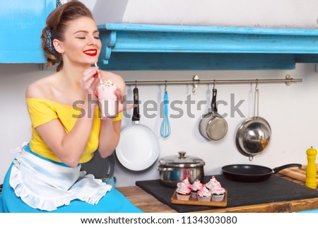 Retro pin up girl woman female housewife wearing colorful top, skirt and white apron holding  sweet strawberry milkshake sitting on the cabinet in the kitchen with utensils and tray with cupcakes. 