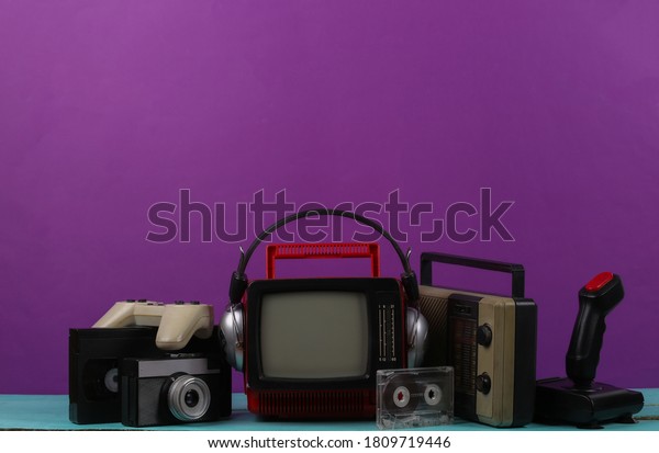 Retro portable\
mini tv set with headphones, radio receiver, joystick, gamepad,\
audio and video cassette, radio receiver on purple background.\
Attributes 80s, retro\
style