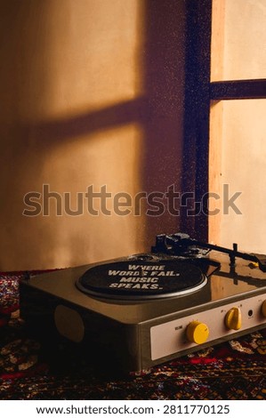 A retro record player with yellow knobs sits on a patterned rug, warm golden light casting window shadows on the wall, vinyl reading 