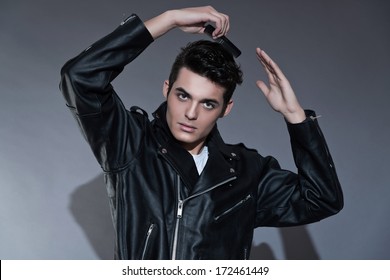 Retro Rock And Roll 50s Fashion Man. Combing His Hair. Wearing Black Leather Jacket. Studio Shot Against Grey.