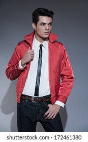 Retro Rock And Roll Fifties Fashion Man With Dark Grease Hair. Wearing Red Jacket And Jeans. Studio Shot Against Grey.