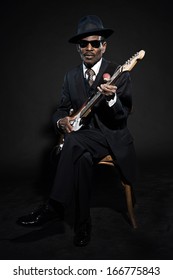 Retro Senior Afro American Blues Man. Wearing Striped Suit With Blue Hat And Black Sunglasses. Playing Electric Guitar.