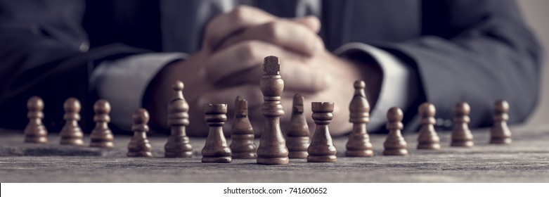 Retro style image of a businessman with clasped hands planning strategy with chess figures on an old wooden table. - Shutterstock ID 741600652