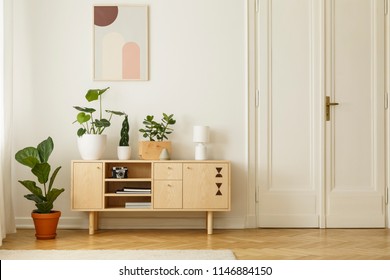 Retro style, wooden sideboard with green plants and a poster on a white wall in a simple apartment interior with herringbone hardwood floor. Real photo.