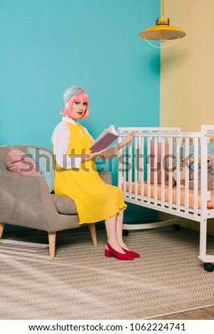 retro styled pregnant pin up woman with pink hair holding book near baby cot in child room