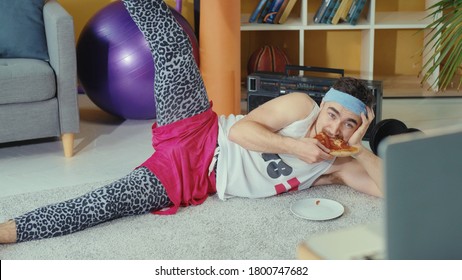 Retro stylish young man jerk eating pizza while doing leg exercises and watching training classes on laptop online. Fitness comedy. Sports and humor concept.