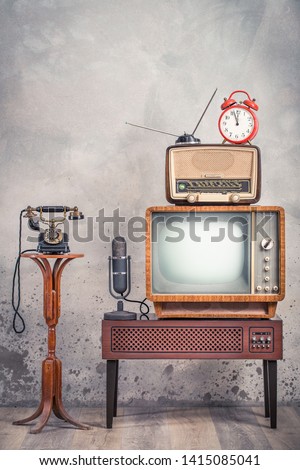 Retro television from 50s, old microphone, radio receiver, orange alarm clock on wooden TV stand and outdated telephone front aged textured concrete wall background. Vintage style filtered photo
