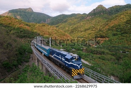 A retro tourist train (Breezy Blue) travels on a viaduct (in South Link Railway Line) over a green lush valley under majestic mountains, in Nanshi, Shihzih Township, Pingtung County, Taiwan