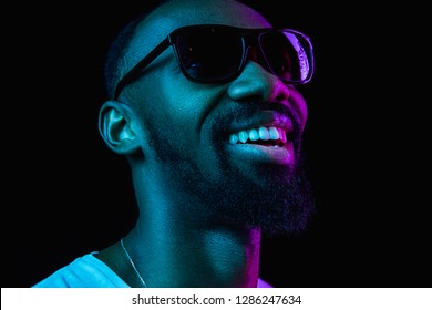 Retro Wave Portrait Of A Young Happy Smiling African Man In Sunglasses At Studio. High Fashion Male Model In Colorful Bright Lights Posing On Black Background. Art Design Concept