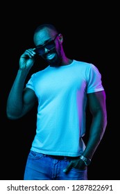 The Retro Wave Or Synth Wave Portrait Of A Young Happy Smiling African Man In Sunglasses At Studio. High Fashion Male Model In Colorful Bright Neon Lights Posing On Black Background. Art Design