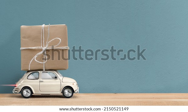 Retro white toy car\
delivering gift box wrapped in kraft paper atop on blue background.\
Copy space