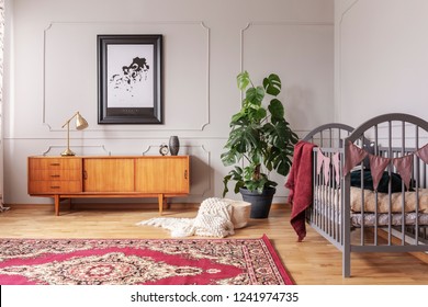 Retro wooden cabinet in grey baby bedroom interior with monster plant in black pot and wooden crib, real photo with poster on the wall