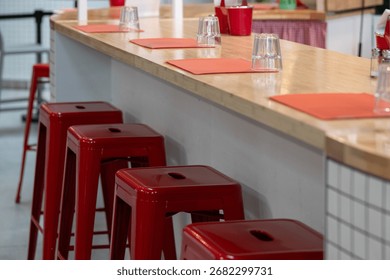 A retro-style diner interior with stackable red metal stools at a white counter topped in cream. Cloth placemats and clear drinking glasses line the counter, evoking a classic, nostalgic charm.
