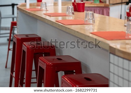 A retro-style diner interior with stackable red metal stools at a white counter topped in cream. Cloth placemats and clear drinking glasses line the counter, evoking a classic, nostalgic charm.