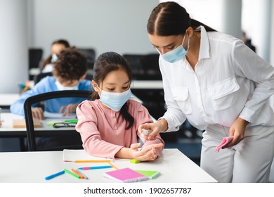 Return Back To School With New Rules. Young Female Teacher In Protective Surgical Face Mask Applying Antibacterial Sanitizer Spray On Asian Pupil's Hands, Working In Classroom At School