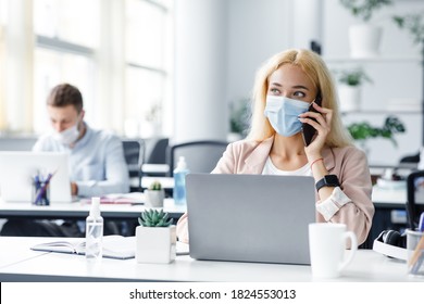 Return To Work After Covid-19 Quarantine. Busy Millennial Woman In Protective Mask Talking On Phone And Typing On Laptop At Workplace With Antiseptic In Modern Office Interior, Free Space